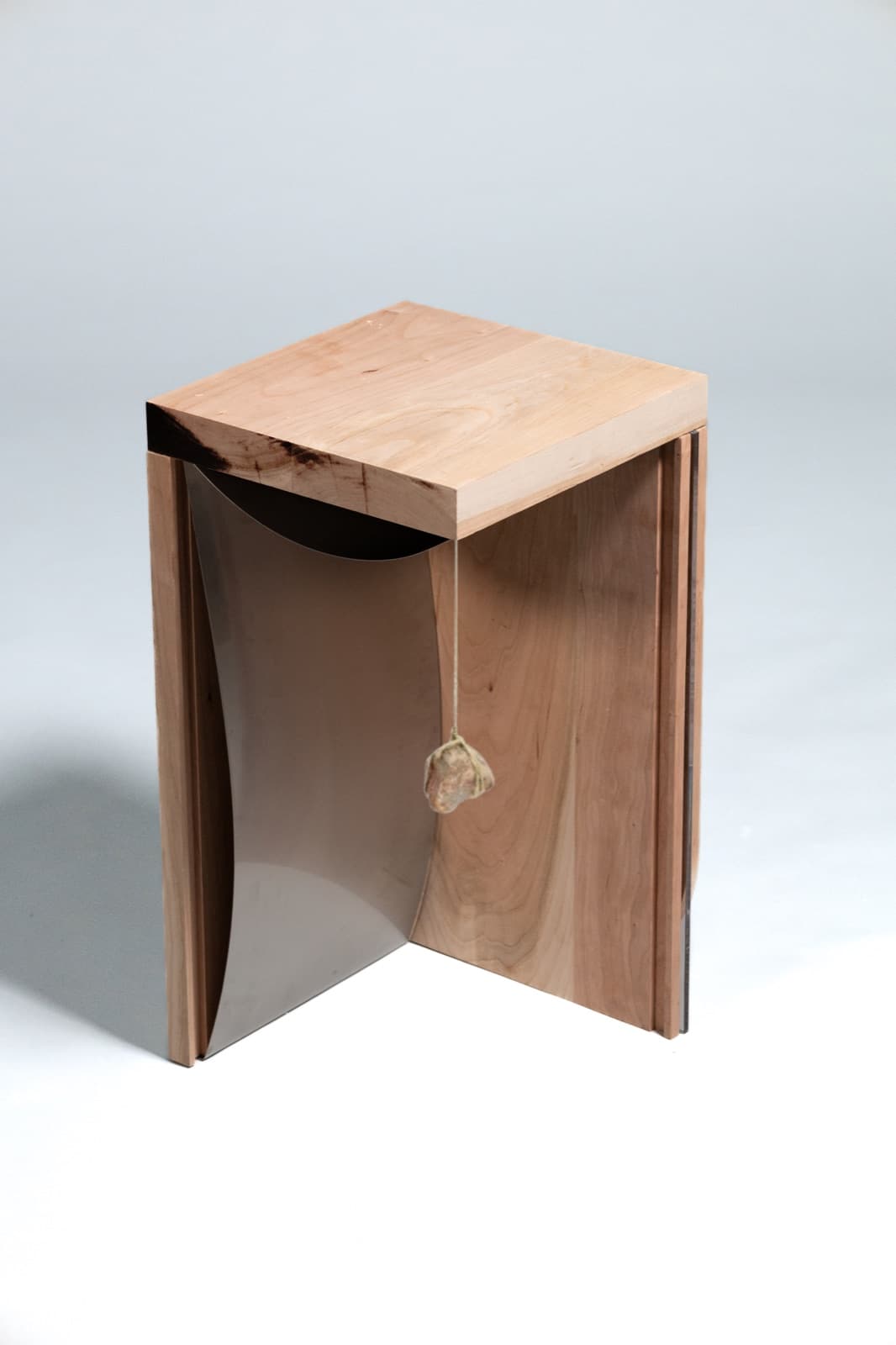 A wooden side table with a curved stainless steel panel and a wrapped stone hanging from a string inside it